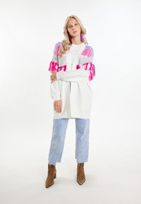 Vit kofta med färgglada stickade paneler i rosa, blått och grått, med fransdetaljer. Matchas med ljusblå jeans och bruna klackboots.