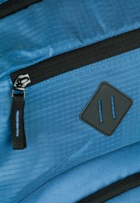 Modrý textilní batoh s texturovaným povrchem, s černým zipem a detaily. Obsahuje černou nášivku ve tvaru diamantu.