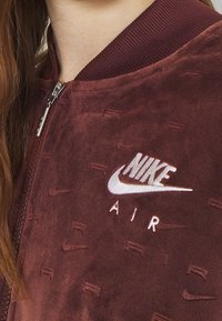 Burgundská bomber bunda se zipem z jemné a strukturální látky. Obsahuje bílou vyšitou nášivku "Nike Air" a žebrovaný límec.