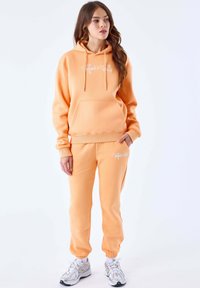 Peachfarvet hoodie og sweatpants sæt, lavet af fleece materiale. Har en frontlomme, snører og hvide logo accenter.