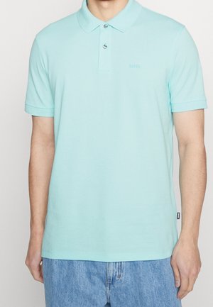 Homme portant un polo bleu clair à manches courtes avec deux boutons et un jean bleu clair, debout devant un fond uni.