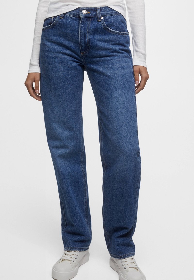 Vaqueros de denim azul con corte recto, cinco bolsillos y cierre de botón. Combinados con una camisa blanca de manga larga y zapatillas blancas.