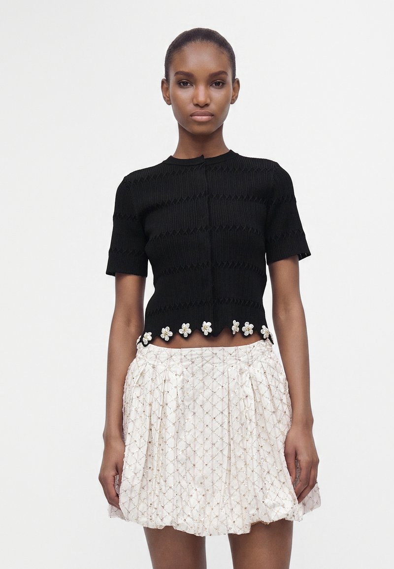 Femme portant un haut noir à manches courtes avec des détails floraux à l’ourlet et une mini-jupe blanche texturée avec un motif en losange.