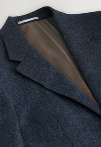 Marineblaues Wollblazer mit Reverskragen, strukturierter Außenseite und glattem braunem Innenfutter mit Fischgrätmuster.