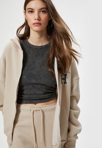 Beige zip-up hoodie med en mørkegrå ribbet top under. Beige sweatpants med snøre. Model med langt hår og hoops øreringe.