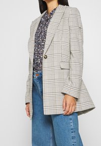 Ternet blazer i beige og sort, med en enkelt knap, skarpe slag og en let oversized pasform. Sættes sammen med en blomstret bluse og brede jeans.