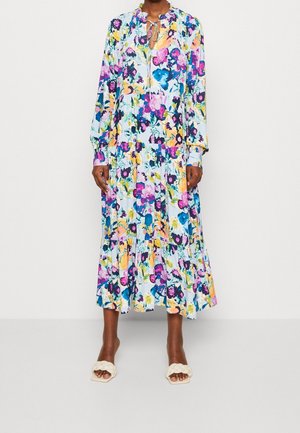 Langärmliges, knöchellanges Kleid mit buntem Blumenmuster und Rüschenkragen, kombiniert mit weißen Sandalen mit geflochtenem Riemen vor hellem Hintergrund.