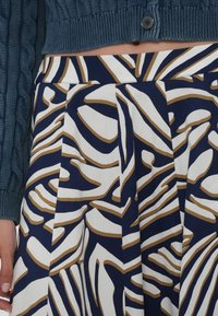 Mornarica suknja s apstraktnim uzorkom u bijeloj i zlatnoj boji, sa plisiranim detaljima i dugim tamno tirkiznim pletenim puloverom na dugmad. Glatka tkanina, strukturiran dizajn.