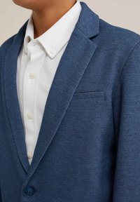 Blazer blu con texture, colletto a rotondo, taschino sul petto e chiusura con bottoni. Indossato sopra una camicia bianca con colletto e bottoni visibili.