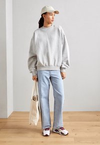 Jasnoszara bluza o luźnym kroju, niebieskie dżinsy i wielokolorowe sneakersy. Beżowa czapka i kremowa torba na ramię dopełniają stylizację.