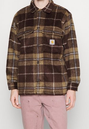 Mann trägt eine braun-gelb karierte Fleecepulloverjacke mit aufgesetzten Brusttaschen und hellrosa Hose vor einfarbigem Hintergrund.