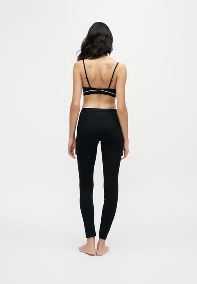 Femme debout pieds nus, tournée de dos, portant un soutien-gorge noir et des leggings, avec deux tatouages d'yeux sur le haut du dos.