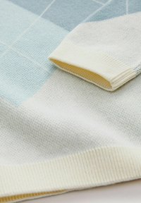 Sweater bleu clair et crème avec un tricot texturé, présentant un motif à carreaux et des poignets côtelés. Les poignets sont crème avec une subtile touche de jaune.