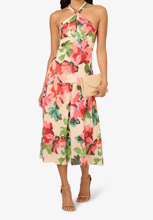 Kobieta w kwiecistej sukience na ramiączkach z odkrytymi ramionami, trzymająca beżową kopertówkę, w połączeniu z cielistymi szpilkami na kostkę.