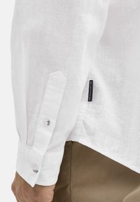 Witte linnen blouse met lange mouwen, knoopmanchetten en een zijlabel. De stof heeft een gestructureerde, ademende uitstraling en gevoel.