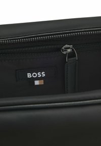 Interno di una borsa in tessuto nero con tasca con zip, etichetta BOSS con tre strisce orizzontali in nero, bianco e marrone. Texture liscia.