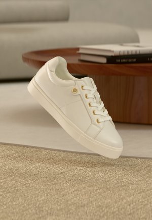 White leather sneaker with gold eyelets and logo pin, placed near a wooden round table with stacked books in a modern living room.