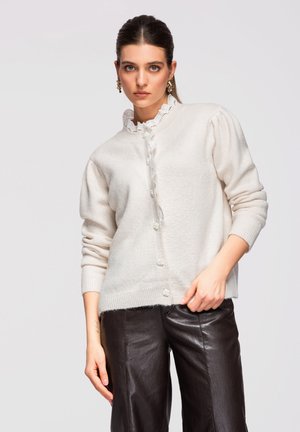 Donna che indossa un cardigan bianco con bottoni e scollo in pizzo e pantaloni in pelle neri su uno sfondo chiaro e semplice.