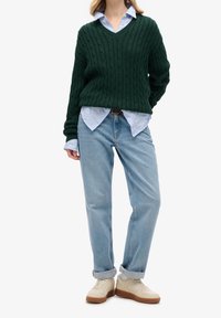 Woman wearing a dark green cable-knit V-neck sweater over a light blue shirt, light blue jeans with cuffs, and beige sneakers with white socks.