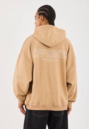 Mann med flettet hår iført beige hettegenser med hetta oppe, tekst "OFF LIMITS DANGEROUS AREA BERLIN 2001" på ryggen, og mørke bukser.