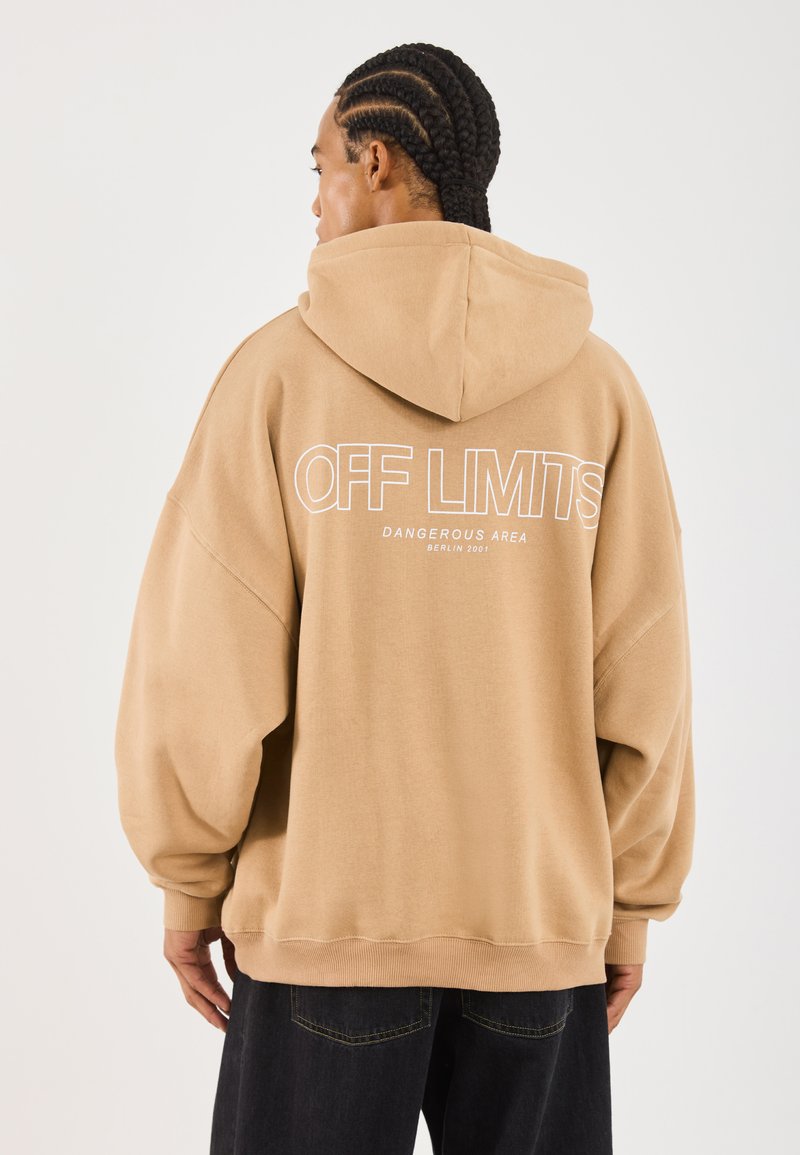 Hombre con cabello trenzado que lleva una sudadera beige con capucha levantada, con el texto "ZONA PELIGROSA RESTRINGIDA BERLÍN 2001" en la parte de atrás, y pantalones oscuros.