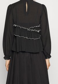 Svart topp med långa ärmar, med en volangig, lager-på-lager design med vit kantning. Nyckelhålsdetalj i ryggen och strukturerad tyg.