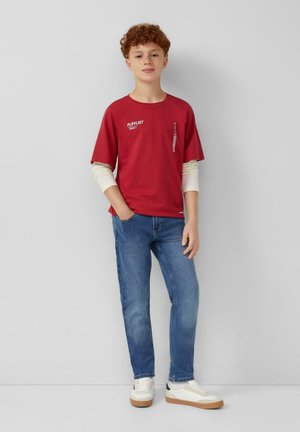 Chłopiec z rudymi, kręconymi włosami, ubrany w warstwowe czerwono-kremowe długie rękawy, niebieskie dżinsy i białe trampki, stojący przy szarej ścianie.