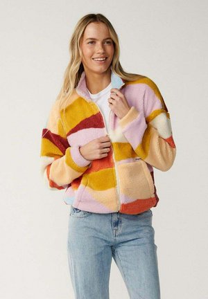 Kvinde smiler og bærer en flerfarvet fleecejakke med pink, gul, hvid, rød og lyseblå abstrakte mønstre og lyseblå jeans.