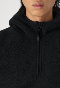 Černý fleecový pulovr s polovičním zipem. Texturovaný materiál, kapuce a hladký zip kontrastují s látkou.