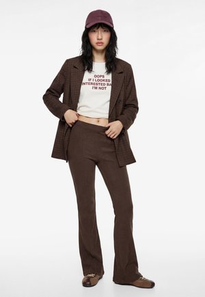 Frau trägt ein braunes kariertes Blazer und Hose, weißes kurzes T-Shirt mit Schrift, dunkelroten Cap und braune Schuhe, steht vor weißem Hintergrund.