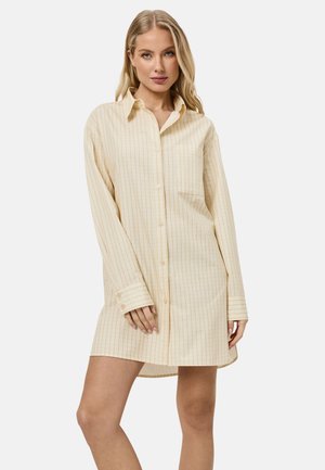 Mujer rubia vestida con un vestido camisa de manga larga a cuadros en crema y naranja claro, con botones delanteros y un bolsillo en el pecho.