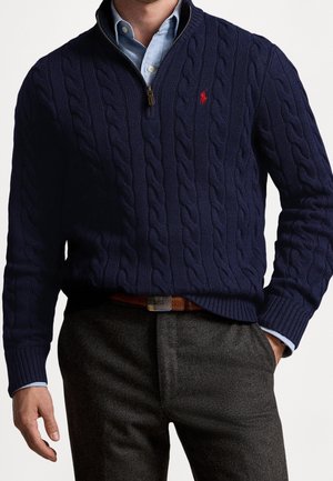 Granatowy sweter w splot warkoczowy z kołnierzem na zamek, z czerwonym logo. Nosi się go na jasnoniebieską koszulę z kołnierzykiem, w połączeniu z ciemnymi teksturowanymi spodniami.