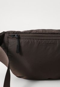 Brown nylon crossbody bag with a textured finish, featuring a zippered pocket, adjustable strap, and ARKK Copenhagen branding.
