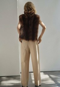 Bruine bonten vest met een hoge kraag, gecombineerd met beige wijde broekspijpen. Gladde textuur en minimalistisch ontwerp, geplaatst tegen een lichte achtergrond.