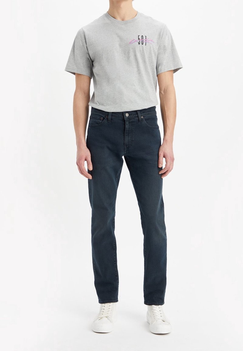 Grå bomullströja med en liten grafisk design på bröstet, ihopparad med mörkblå jeans och vita sneakers. Enkelt mönster, avslappnad passform.