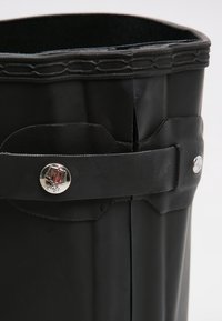 Bota de goma negra con un diseño cilíndrico alto. Presenta una correa plana con un cierre de botón plateado y un borde superior texturizado.