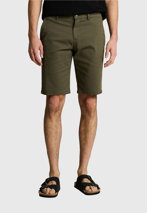 Mężczyzna ubrany w oliwkowozielone krótkie spodnie do kolan i czarne sandały z klamrą, stojący na jednolitym tle.