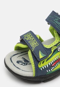 Blå og grønne sandaler med dinosaur-tema, som har justerbare stropper, teksturerte såler og en trykt fotseng med fossildesign.