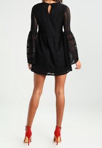 Kvinna som bär en svart spetskortklänning med långa trumpetärmar och röda högklackade ankelsandaletter, visad bakifrån.