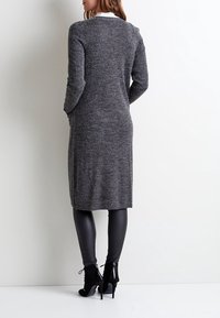 Lång, grå stickad kofta med en strukturerad yta, ribbad nederkant och långa ärmar, i kombination med svarta leggings och högklackade ankelstövlar.