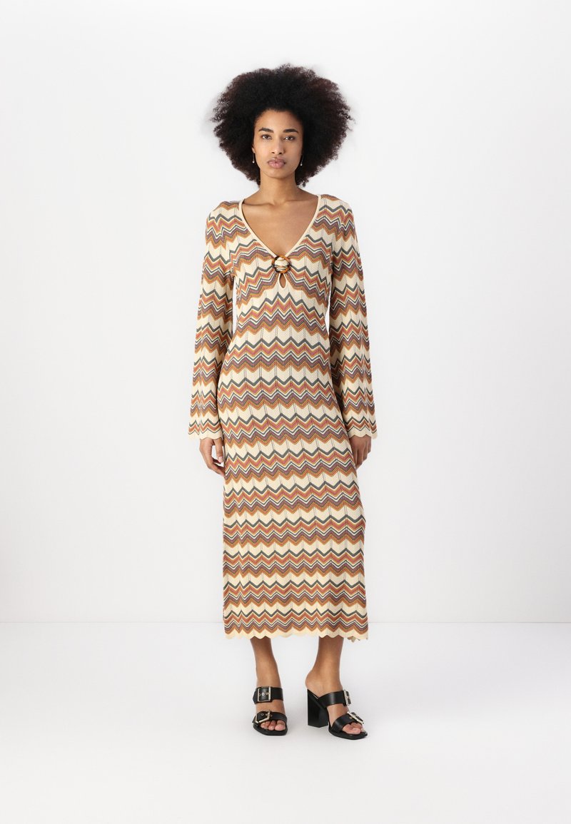 Robe en tricot à manches longues avec un col en V, ornée d'un motif zigzag en orange, marron et crème, et d'une fermeture décorative au niveau du col.