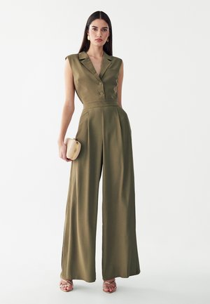 Vrouw draagt een mouwloze olijfgroene jumpsuit met wijde pijpen en knopen, houdt een kleine beige clutch vast en draagt open teenhakken.