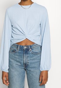 Person som bär en ljusblå långärmad topp med vridning fram, kort modell, och högmidjade blå jeans, samt ett tunt kedjehalsband.