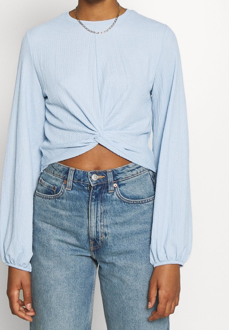 Person som bär en ljusblå långärmad topp med vridning fram, kort modell, och högmidjade blå jeans, samt ett tunt kedjehalsband.