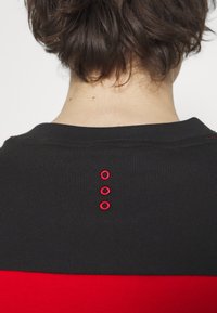 Suéter de punto negro con una franja horizontal roja en la parte inferior y tres círculos rojos bordados a lo largo del cuello trasero.