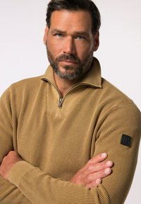 Brauner Strickpullover mit einem halben Reißverschlusskragen, strukturiertem Muster und einem schwarzen Logo-Patch auf dem linken Ärmel. Arme vor der Brust verschränkt.