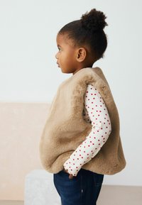 Beige imiterad pälsväst som bärs över en långärmad vit skjorta med röda prickar; blå jeans; barns profilvy med naturligt hår.