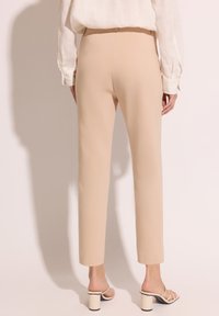 Beżowe spodni z dopasowanym krojem, gładką teksturą i długością 7/8, w połączeniu z jasnymi sandałami na obcasie na neutralnym tle.