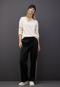 Maglione beige, pantaloni neri con strisce bianche ai lati e sneakers bianche. Tessuti morbidi, vestibilità rilassata e vita con cordino. Berretto nero.
