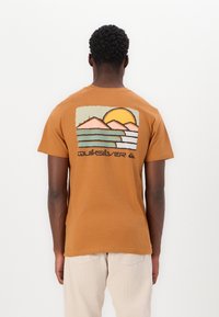 Brun bomull T-skjorte med et fargerikt fjell- og solgrafikk på baksiden, med "Quiksilver" tekst i en dristig, svart skrift.